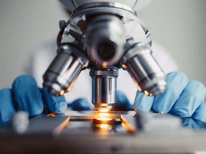 Close up view of someone in blue gloves using a microscope with light on examining of test sample